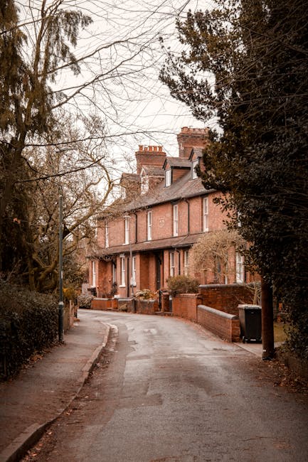 A narrow residential street lined with brick terraced houses in Hadley Wood, surrounded by tall, leafless trees and overcast skies. The pavement runs along the front of the houses, with a slight bend in the road leading to the property at the end. Although no furniture or moving equipment is visible, the scene suggests a typical location for home relocation activities. Man with Van Hadley Wood specializes in removals and moving services, including navigating tight access roads like this one for efficient furniture transport and packing processes. The muted lighting and quiet environment reflect a calm setting suitable for house removals, with the street's constrained width highlighting the need for careful planning and specialized access solutions during a house move.