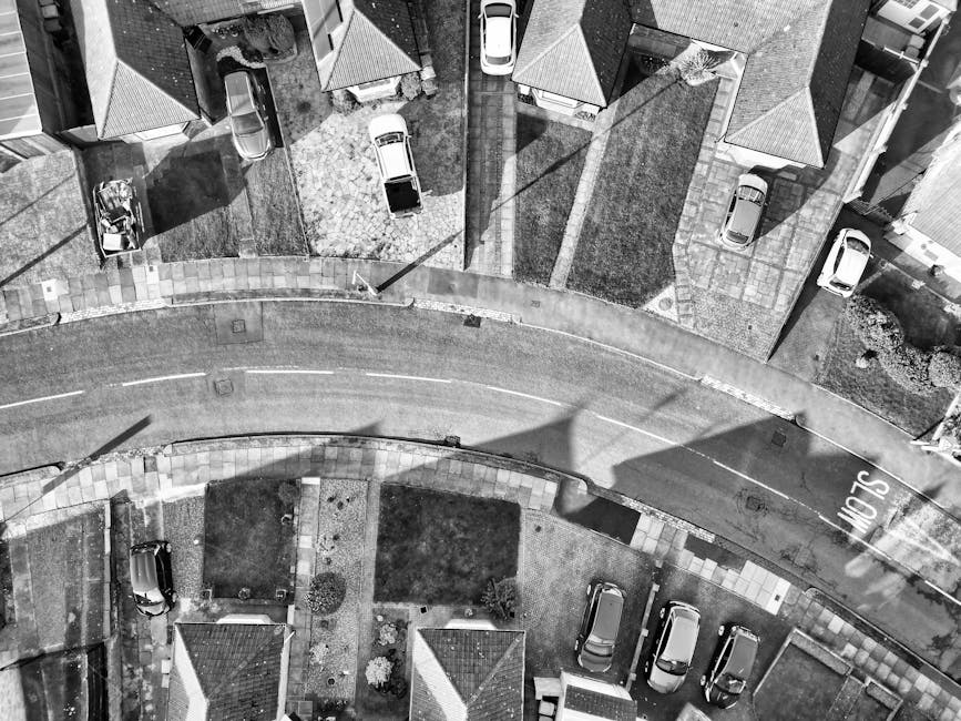 An aerial black-and-white photograph showing a residential street during a home relocation process. Several houses with pitched roofs and small front gardens are arranged along a curved road with parked cars on each side. In the foreground, there is a driveway with packing boxes, some wrapped in plastic and bubble wrap, along with furniture covered in protective blankets. A person is seen lifting a large cardboard box near the entrance of a house, suggesting active packing or loading. A moving trolley or dolly is positioned nearby, aiding the transport of belongings from the property to the waiting van, which is not visible in this shot. The scene is well-lit with natural daylight, and the organized layout indicates a professional removals service, such as Man with Van Hadley Wood, managing the furniture and packing materials during the house removal or home relocation process. No other vehicles are in motion, emphasizing a controlled, careful loading environment characteristic of moving logistics aimed at preventing driveway fines and ensuring safe parking for EN4 removals.