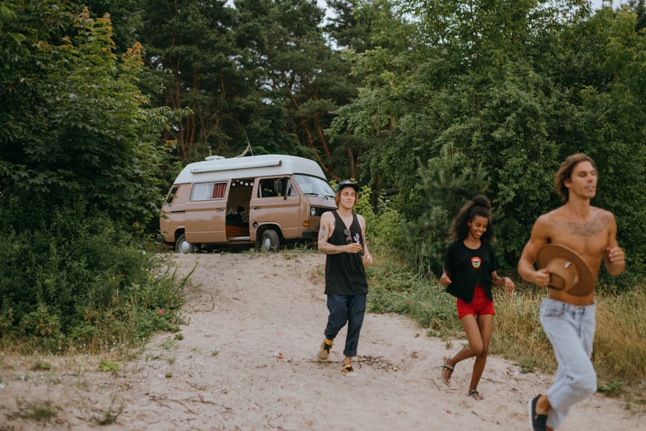 A group of three young adults, two men and one woman, walking along a dirt path surrounded by lush green trees and shrubs during daylight. The man on the right is shirtless, has long hair, tattoos on his chest and arms, and is holding a brown hat while jogging. The woman in the middle has curly hair tied in a bun, wears a black jacket over a T-shirt, red shorts, and beige sandals, and appears to be smiling. The man on the left has tattoos on his arms, wears a black sleeveless vest, dark pants, and sneakers, and is holding what looks like a tumbler. In the background, there is a beige camper van parked on the path, partially hidden by the dense foliage, with its side door open. The van is a typical vehicle used in home relocations for transporting furniture and packed boxes, and the environment suggests an outdoor loading or unloading area near a wooded location, with natural lighting indicating daytime. Man with Van Hadley Wood periodically supported by [COMPANY_NAME] for professional removals, ensuring efficient furniture transport and packing during home moves near Hadley Wood Station.