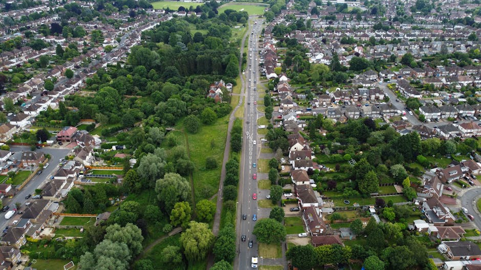 An aerial view of a residential suburb showing a long, straight street with vehicles parked along both sides, flanked by well-maintained houses with gardens and driveways. To the left of the road, there is a large green park area with dense trees, grass, and paved pathways. On the right side, houses with various sizes and designs have landscaped gardens, some with fences and hedges. The street appears busy with cars traveling in both directions, illustrating typical urban traffic. The lighting suggests daytime, with clear weather and natural light illuminating the scene. This image highlights a suburban environment suitable for house removals, with visible potential loading areas near the property driveways, and overall depicts the setting where furniture transport and home relocation operations could take place. Man with Van Hadley Wood may use such locations when planning moving services in the Hadley Wood area, as indicated by the webpage about Enfield Council removals permits.