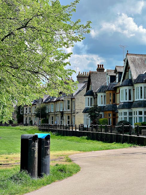 A row of traditional terraced houses in Hadley Wood with brick facades and bay windows, situated along a quiet residential street. In the foreground, two black wheelie bins for household disposal are positioned on a grassy verge next to a paved footpath. A large tree with bright green leaves partially shades the scene on the left side, while a clear blue sky with some white clouds is visible overhead. The image depicts a peaceful suburban setting typical of home relocation and moving logistics areas, with the environment suitable for residential removals and the transportation of household items by professionals such as Man with Van Hadley Wood, who may operate in areas emphasizing household disposal rules and house moves.