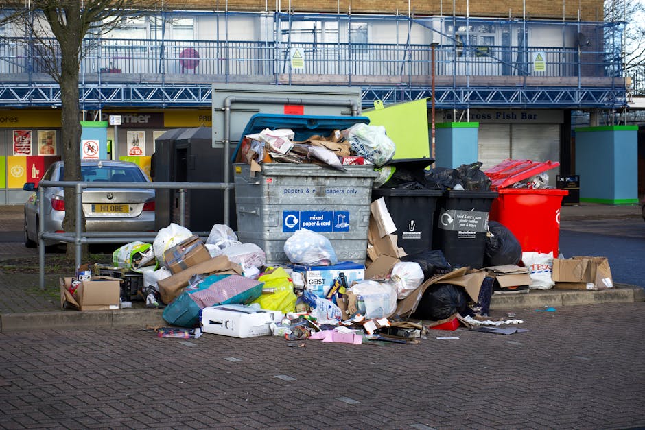 An outdoor scene showing multiple overflowing waste and recycling bins, including a large grey mixed paper and cardboard bin, a black general waste bin, and a red recycling bin, all positioned on a paved area beside a pavement. Surrounding the containers are scattered cardboard boxes, plastic bags filled with rubbish, paper packaging, and other waste materials spilling onto the ground. In the background, there is a building with a yellow storefront that appears to be a fish bar, along with a parked silver car in front of the bins, and a blue scaffolding structure covering part of the building. The scene is illuminated by natural daylight, with leafless trees partially visible on the left side, indicating an urban environment where household waste collection or disposal may be relevant to home relocation and packing processes. Man with Van Hadley Wood regularly handles such waste management during house removals, especially when adhering to local disposal regulations in Enfield and Hadley Wood.