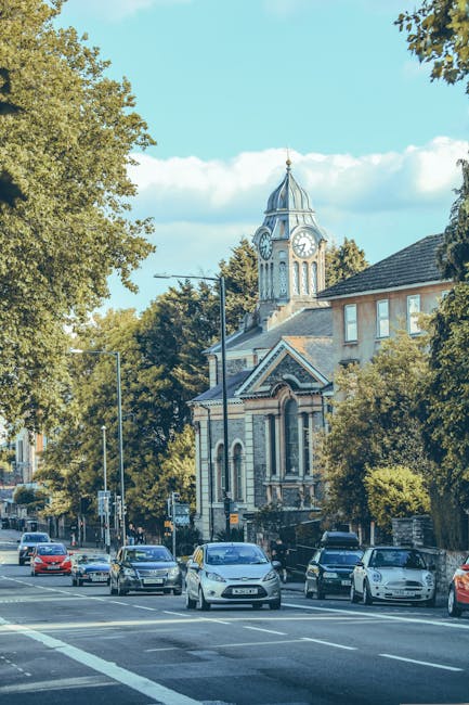An aerial view of a small town in Hadley Wood during daytime, showing a historic stone church surrounded by mature trees with green, yellow, and orange foliage. The church is situated near a curved ro