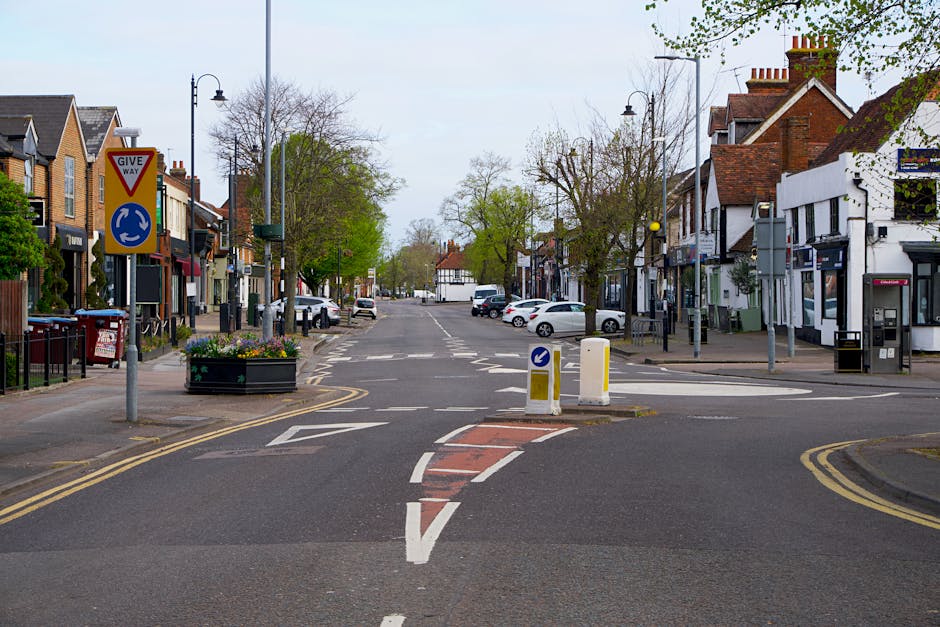 A view of a quiet residential street in Hadley Wood, showing a crossroad with white painted lines and traffic bollards in the center. On the left side, a 'Give Way' sign and a blue roundabout sign are visible alongside flower planters and parked cars. The street is lined with semi-detached houses featuring brick facades, pitched roofs, and small front gardens, some with trees and shrubs. On the right side, there are additional houses, a payphone booth, and street lamps illuminating the scene. The sky is overcast, and the overall environment appears calm and suitable for a home relocation or furniture transport operation, which [COMPANY_NAME] can facilitate through professional moving and packing services. The scene suggests a typical urban neighbourhood prepared for the logistical process of loading and unloading household items during a house removal.